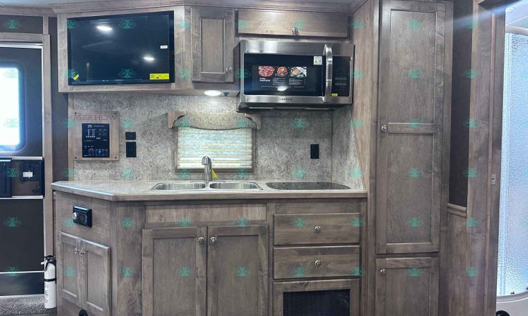 Kitchen interior of a travel trailer with wood cabinets, stainless microwave, sink, and window shade above the counter.
