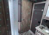 Interior view of an RV bathroom with a curved glass shower enclosure on the left, wood-tone cabinets, and a small sink with a countertop on the right.