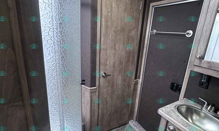 Interior view of an RV bathroom with a curved glass shower enclosure on the left, wood-tone cabinets, and a small sink with a countertop on the right.