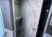 Open walk-in closet in a trailer, showing a hanging rod, shelf, and small bench with gray wood finish panels nearby.