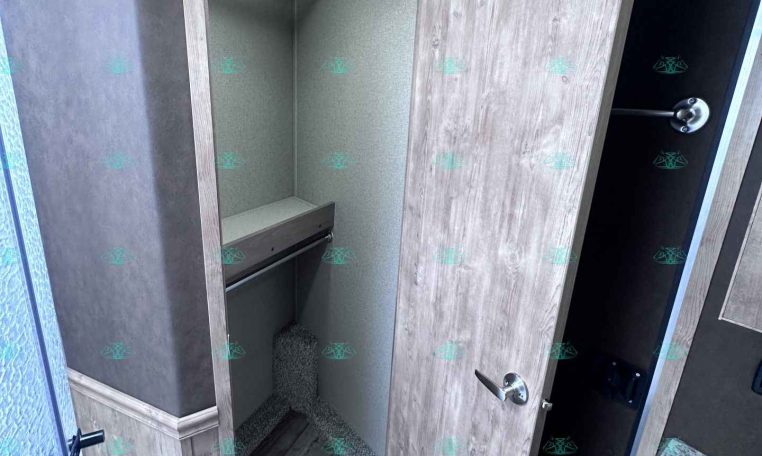Open walk-in closet in a trailer, showing a hanging rod, shelf, and small bench with gray wood finish panels nearby.