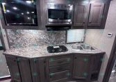 RV kitchen with dark wood cabinets, built-in microwave, stovetop, and double sink on granite counters in a compact trailer galley.