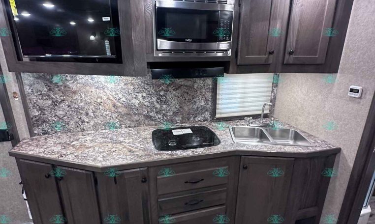 RV kitchen with dark wood cabinets, built-in microwave, stovetop, and double sink on granite counters in a compact trailer galley.