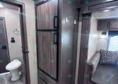 Interior of an RV showing dark wood cabinets, a tall fridge and drawers, with a small bathroom and toilet on the left and a seating area with a patterned cushion on the right.