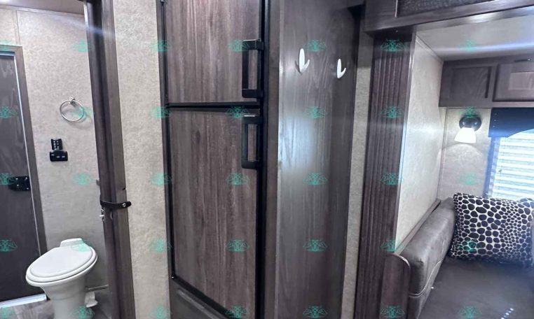 Interior of an RV showing dark wood cabinets, a tall fridge and drawers, with a small bathroom and toilet on the left and a seating area with a patterned cushion on the right.