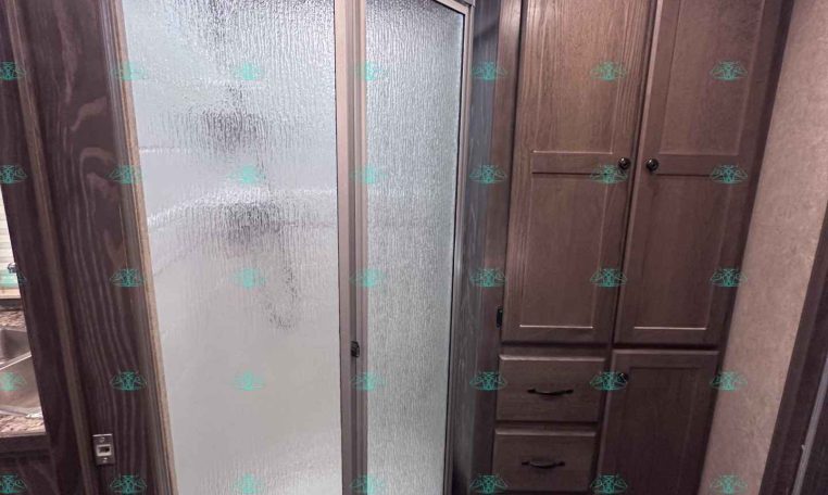 Bathroom interior of a travel trailer featuring a frosted glass shower enclosure on the left and a tall wooden cabinet unit on the right, with a ceiling vent and recessed light above.