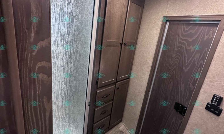 Small trailer bathroom with a frosted glass shower door, wooden cabinets, and a toilet in the foreground.
