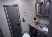 Small bathroom in a trailer with a toilet, stainless sink, granite countertop, and dark wood vanity with a mirrored cabinet above. A vase of sunflowers sits on the counter. Dim overhead lighting and a door to the left.