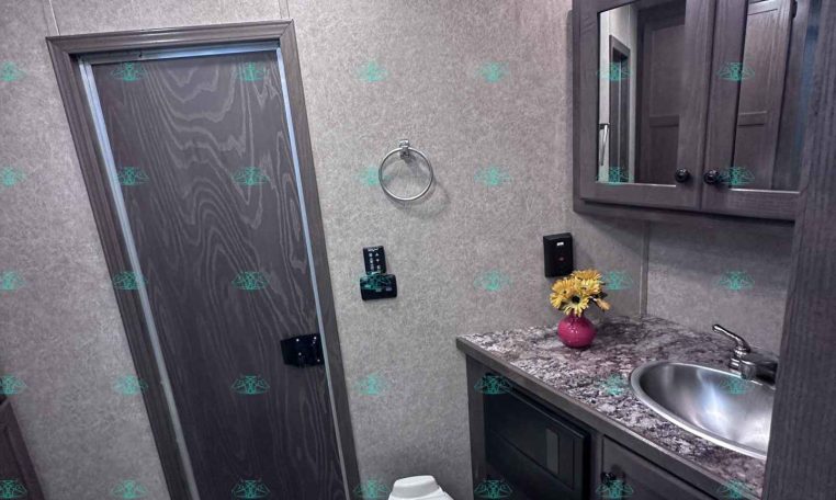 Small bathroom in a trailer with a toilet, stainless sink, granite countertop, and dark wood vanity with a mirrored cabinet above. A vase of sunflowers sits on the counter. Dim overhead lighting and a door to the left.