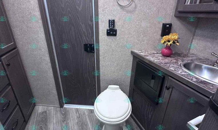 RV bathroom with dark wood cabinets, granite countertop, sink, and a toilet; pink vase with yellow flowers on the counter nearby.