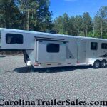Silver horse trailer parked on a gravel lot with a forest of tall trees and blue sky in the background, branding on the side visible.