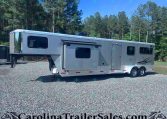 Silver horse trailer parked on a gravel lot with a forest of tall trees and blue sky in the background, branding on the side visible.