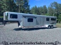 Silver horse trailer parked on a gravel lot with a forest of tall trees and blue sky in the background, branding on the side visible.