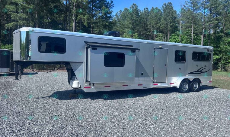 Silver horse trailer parked on a gravel lot with a forest of tall trees and blue sky in the background, branding on the side visible.