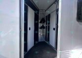 Open trailer door revealing a narrow aluminum trailer interior with white cabinetry along the walls and a small metal rack at the far end.