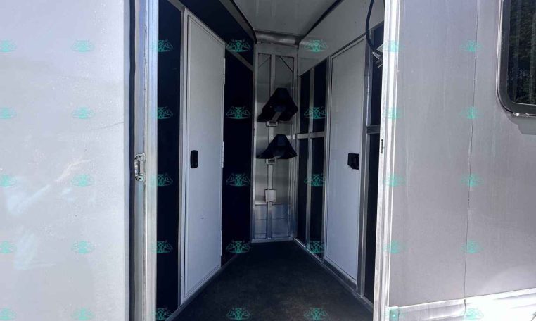 Open trailer door revealing a narrow aluminum trailer interior with white cabinetry along the walls and a small metal rack at the far end.