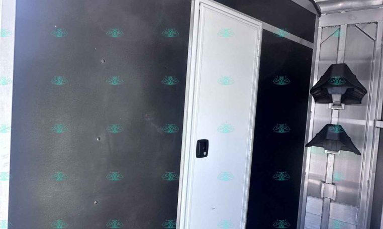 Interior of a horse trailer showing black wall panels, a white cabinet, and metal framing with a row of black helmet racks on the right side.