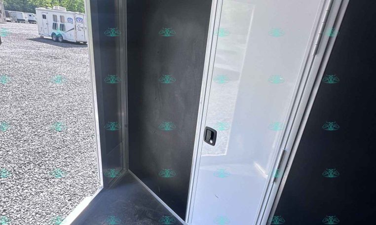 Interior of a black trailer with silver trim and a white door, open to a gravel lot and a neighboring horse trailer outside.