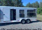 Silver aluminum horse trailer with open side door and barred windows, parked on a gravel lot with trees in the background (NTS logo visible).
