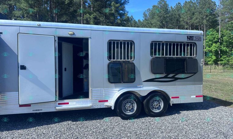 Silver aluminum horse trailer with open side door and barred windows, parked on a gravel lot with trees in the background (NTS logo visible).