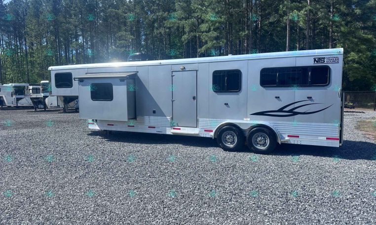 Silver horse trailer parked on gravel with trees in the background; features a side door, multiple windows, and a decorative graphic near the rear.