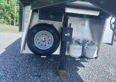 Trailer hitch area with spare tire mounted on back, two propane tanks, and wheel jack in a gravel lot.