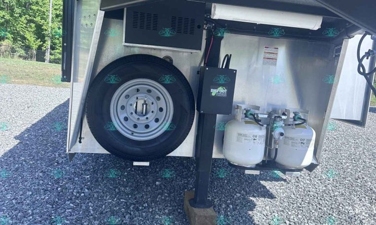 Trailer hitch area with spare tire mounted on back, two propane tanks, and wheel jack in a gravel lot.