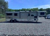 Wide view of a large white fifth-wheel RV parked on a gravel lot with trees in the background, under a clear blue sky.