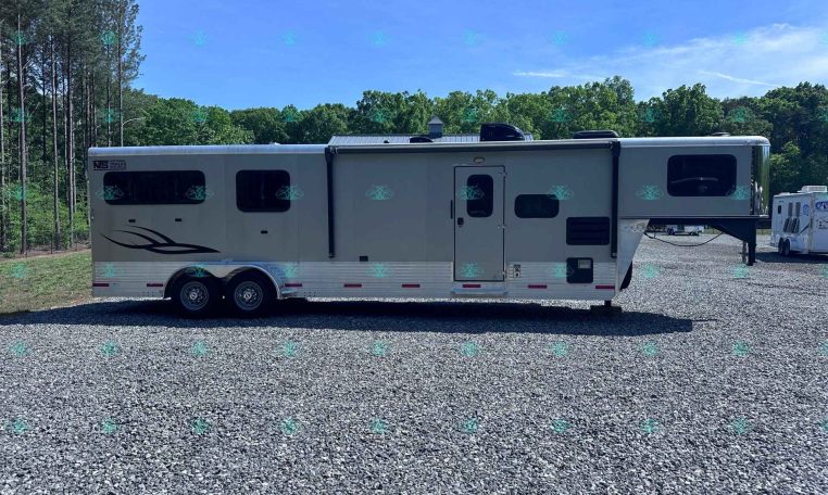 Wide view of a large white fifth-wheel RV parked on a gravel lot with trees in the background, under a clear blue sky.