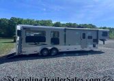Large beige horse trailer with open rear ramp, parked on a gravel lot with green trees and blue sky in the background.