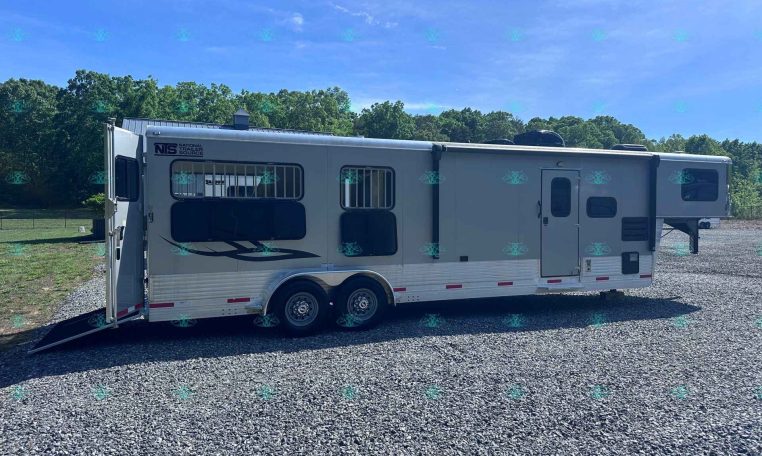 Large beige horse trailer with open rear ramp, parked on a gravel lot with green trees and blue sky in the background.