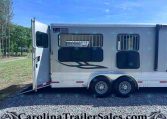Silver horse trailer with two barred windows and an open side ramp, parked on gravel with a blue sky and trees in the background; logo reads National Trailer Source on the side.