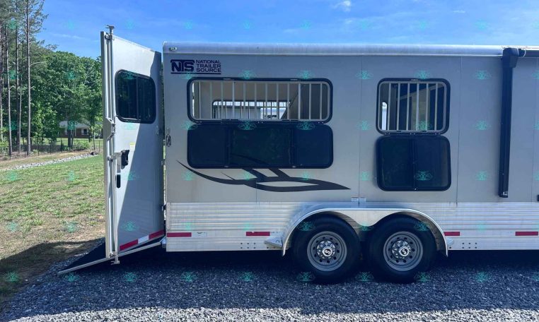 Silver horse trailer with two barred windows and an open side ramp, parked on gravel with a blue sky and trees in the background; logo reads National Trailer Source on the side.