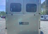 Rear of a light-gray trailer with two rear doors featuring rounded windows and the National Trailer Source logo on the lower half, on a gravel lot.