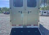 Rear view of a gray enclosed trailer with double doors, windows in each door, and a lowered black loading ramp on a gravel lot. In the background, other trailers are parked.