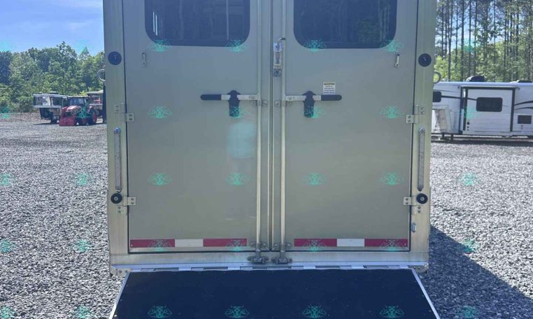 Rear view of a gray enclosed trailer with double doors, windows in each door, and a lowered black loading ramp on a gravel lot. In the background, other trailers are parked.