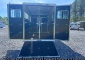 Back of a dark horse trailer with doors open and a lowered ramp, showing stall dividers inside on a gravel lot.