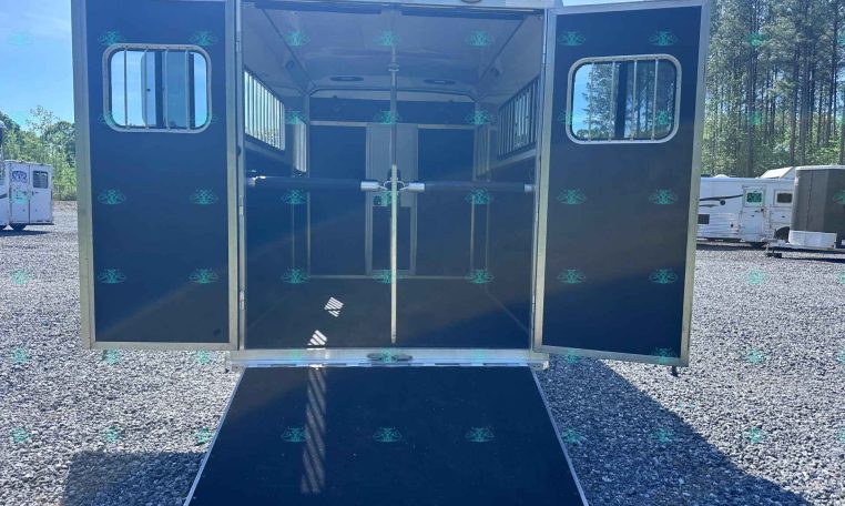 Back of a dark horse trailer with doors open and a lowered ramp, showing stall dividers inside on a gravel lot.