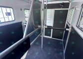 Interior of a horse trailer showing a metal divider gate, black rubber flooring, and metallic walls with windows on the side