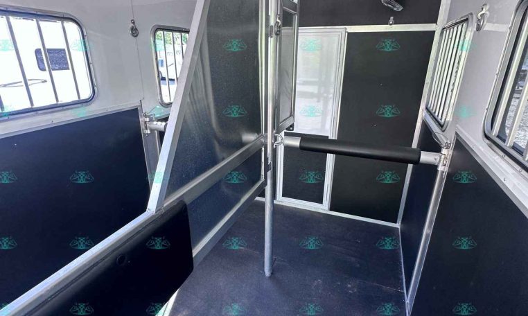 Interior of a horse trailer showing a metal divider gate, black rubber flooring, and metallic walls with windows on the side