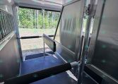 Interior of a silver horse trailer with stalls, open side door, and forest view outside.