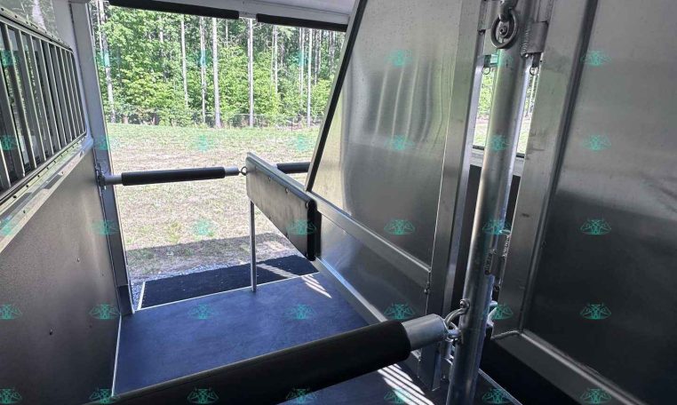 Interior of a silver horse trailer with stalls, open side door, and forest view outside.