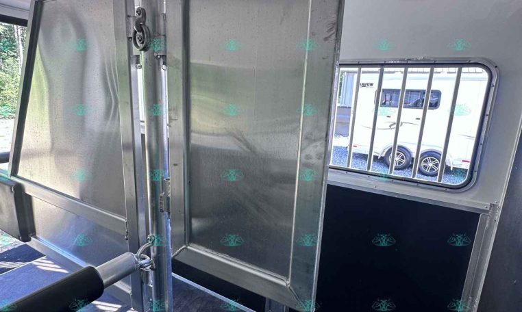 Interior of a metal trailer showing a vertical divider gate and a latching mechanism, with a window and bars visible outside.