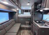 Interior of a compact RV showing a gray sofa on the left, kitchen counter and microwave on the right, and a bed area toward the back with striped bedding at the far end