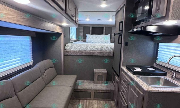Interior of a compact RV showing a gray sofa on the left, kitchen counter and microwave on the right, and a bed area toward the back with striped bedding at the far end