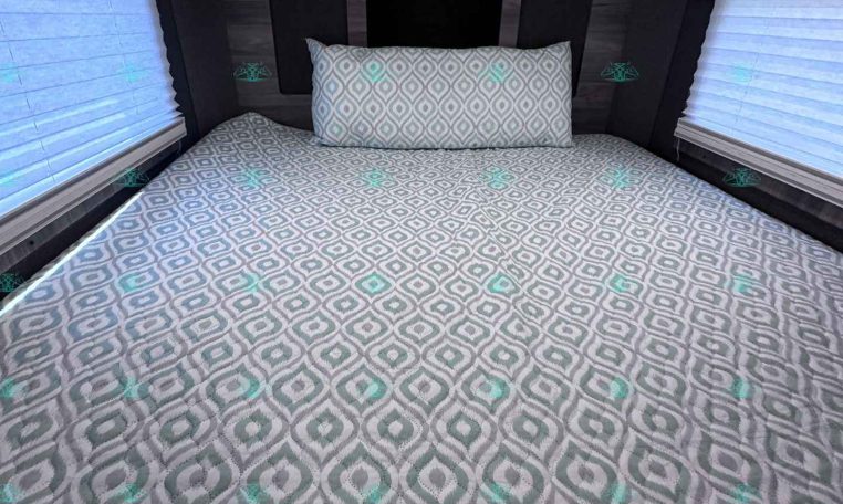 Interior of a trailer bedroom with a bed covered in gray geometric bedding and a matching pillow, flanked by two window blinds and ceiling lights.