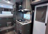 Interior of a modern travel trailer: compact kitchenette with sink, stove, microwave, and upper cabinets beside a doorway and exit sign; bed area visible in background.