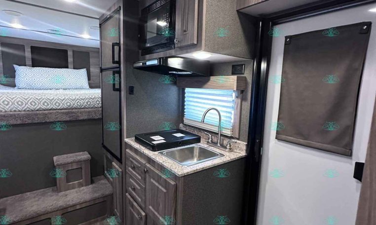 Interior of a modern travel trailer: compact kitchenette with sink, stove, microwave, and upper cabinets beside a doorway and exit sign; bed area visible in background.