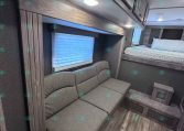 RV living space with gray sectional sofa, overhead cabinets, and a raised bunk bed to the right.