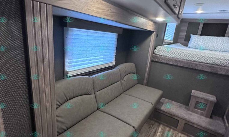 RV living space with gray sectional sofa, overhead cabinets, and a raised bunk bed to the right.